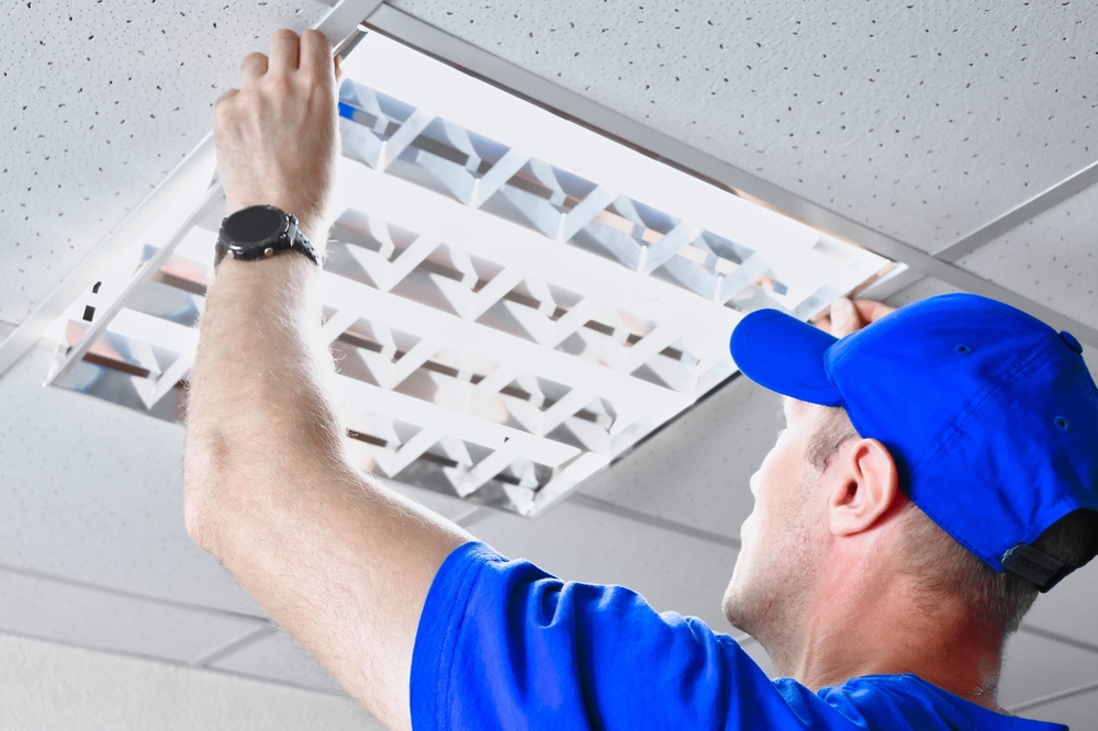 A technician is securing and adjusting a fluorescent light panel in the ceiling of a commercial office, wearing blue work clothes, and ensuring everything is in place for optimal lighting
